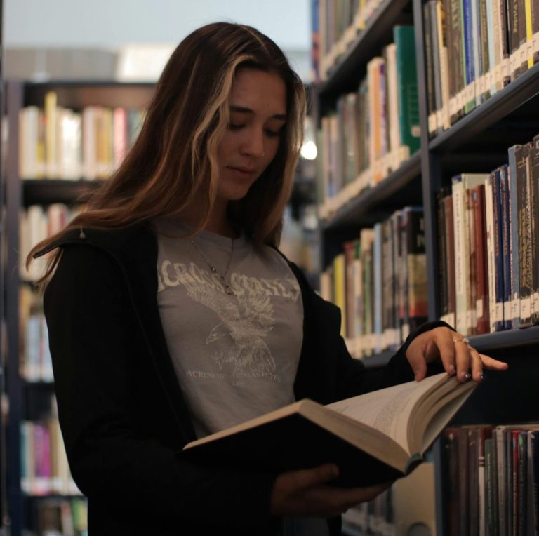 A young woman reading a book in a calm library setting, surrounded by shelves.